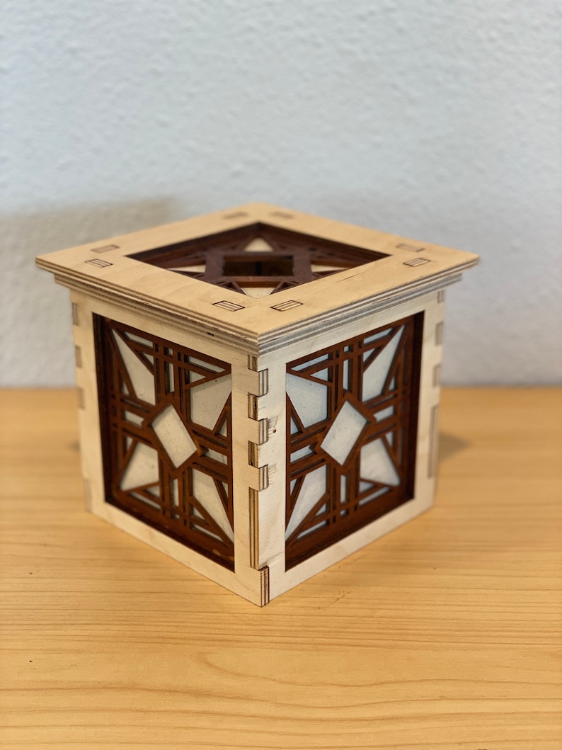 Puede incluir: Una caja de madera con un dise&ntilde;o geom&eacute;trico. La caja est&aacute; hecha de madera clara con detalles en marr&oacute;n oscuro. El dise&ntilde;o es intrincado y presenta una forma de estrella en el centro.