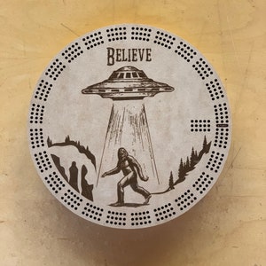 May include: A round cribbage board with a Bigfoot and UFO design. The board features the word "BELIEVE" above the UFO. The board has numerous holes around the perimeter for peg placement and a "START" label.