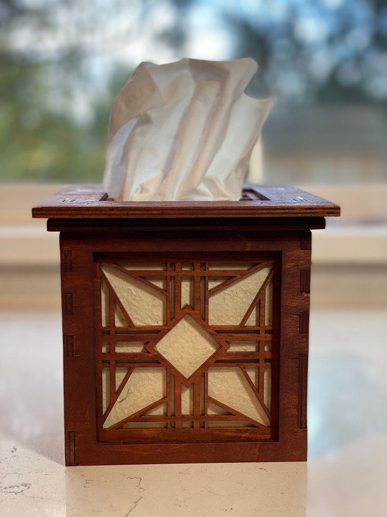 Puede incluir: Una caja de pa&ntilde;uelos de madera con un dise&ntilde;o geom&eacute;trico. La tapa es de madera oscura y tiene una incrustaci&oacute;n de madera clara. La caja de pa&ntilde;uelos est&aacute; sobre una superficie blanca.