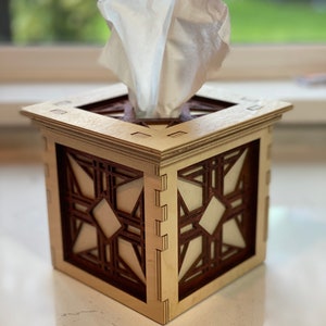 Puede incluir: Un porta pa&ntilde;uelos de madera con un dise&ntilde;o geom&eacute;trico. La caja est&aacute; hecha de madera clara con detalles en marr&oacute;n oscuro. El porta pa&ntilde;uelos est&aacute; sobre una superficie blanca.
