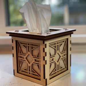 Puede incluir: Un porta pa&ntilde;uelos de madera con un dise&ntilde;o geom&eacute;trico. La caja est&aacute; hecha de madera clara con detalles en madera oscura. El porta pa&ntilde;uelos est&aacute; sobre una encimera clara.