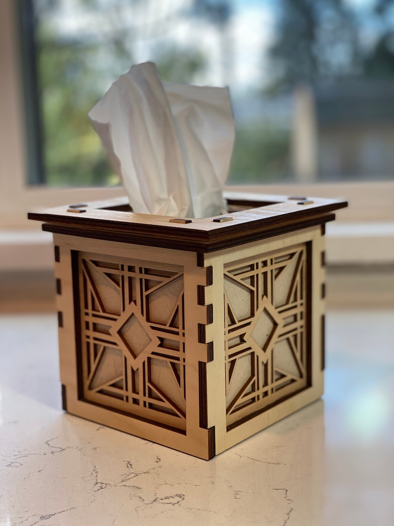 Puede incluir: Un porta pa&ntilde;uelos de madera con un dise&ntilde;o geom&eacute;trico. La caja est&aacute; hecha de madera clara con detalles en madera oscura. El porta pa&ntilde;uelos est&aacute; sobre una encimera clara.