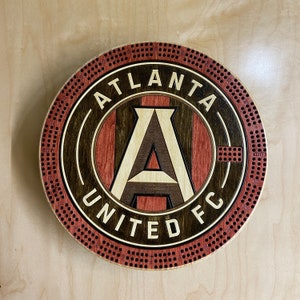 May include: A wooden Atlanta United FC logo with a red and black background and a white and brown letter "A" in the center.