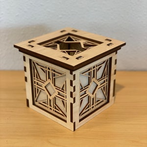 Puede incluir: Una caja de madera con un dise&ntilde;o geom&eacute;trico recortado en los lados. La caja est&aacute; hecha de madera clara con un ribete de madera oscura. La caja est&aacute; sentada sobre una superficie de madera.