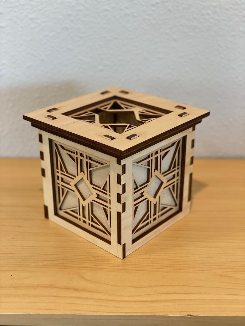 Puede incluir: Una caja de madera con un dise&ntilde;o geom&eacute;trico recortado en los lados. La caja est&aacute; hecha de madera clara con un ribete de madera oscura. La caja est&aacute; sentada sobre una superficie de madera.