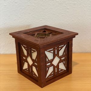Puede incluir: Una caja de madera con un dise&ntilde;o geom&eacute;trico recortado en los lados. La caja est&aacute; te&ntilde;ida de un color marr&oacute;n oscuro y tiene un forro de papel blanco en el interior.