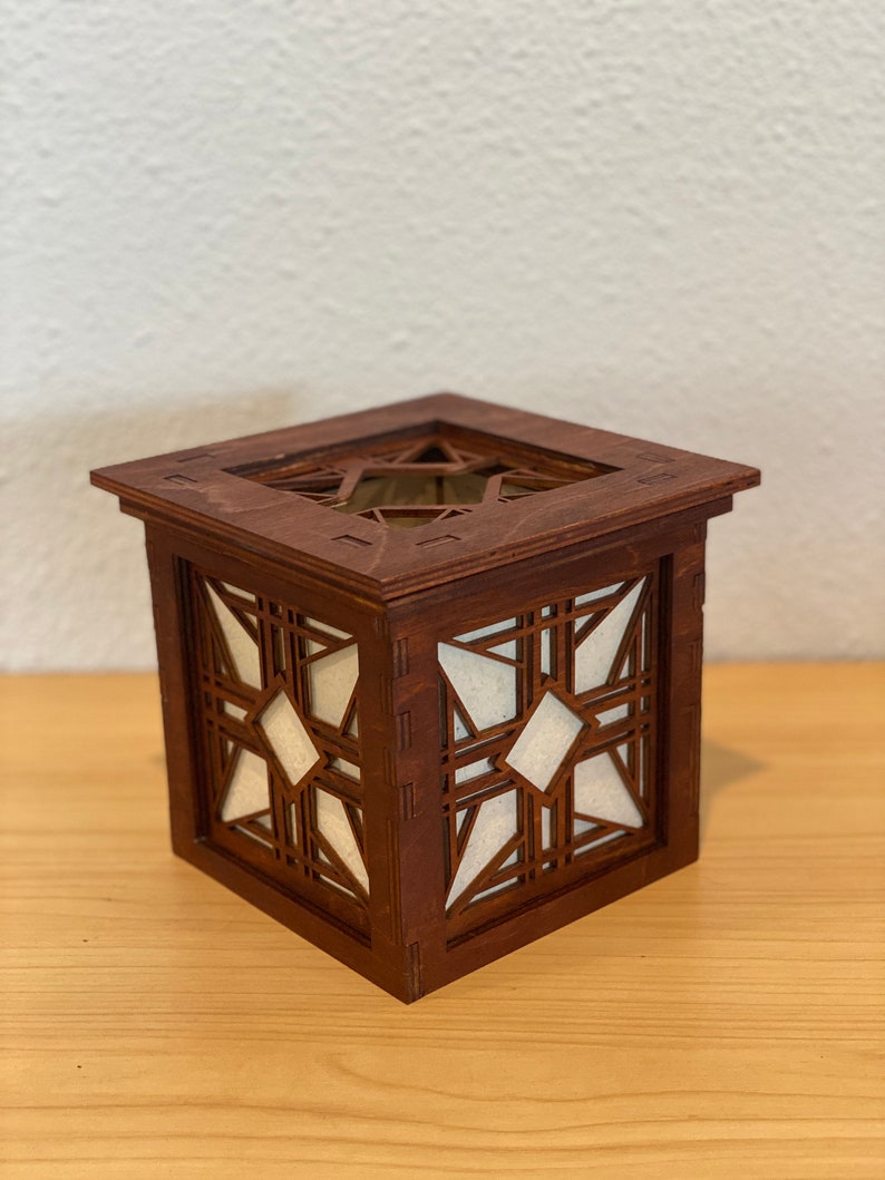 Puede incluir: Una caja de madera con un dise&ntilde;o geom&eacute;trico recortado en los lados. La caja est&aacute; te&ntilde;ida de un color marr&oacute;n oscuro y tiene un forro de papel blanco en el interior.
