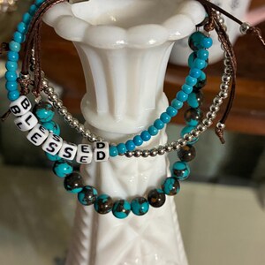 May include: Three beaded bracelets with turquoise and brown beads, and silver accents. One bracelet spells out the word "BLESSED" with white letter beads. The bracelets are displayed on a white decorative vase.