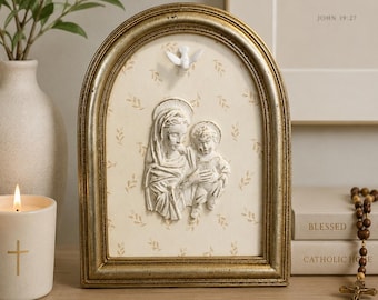 May include: An arched wall plaque featuring a white relief sculpture of a mother and child centered on a cream background with a delicate leaf pattern. The piece is encased in a distressed gold frame and includes a small white dove at the top.
