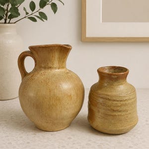 May include: Two handcrafted ceramic vases in earthy tones. The larger vase has a handle and a spout, while the smaller one is a rounded shape. Both have a textured surface and a rustic aesthetic, perfect for home decor.