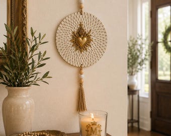 May include: A circular wall hanging featuring a textured cream-colored disc with a central gold-toned Sacred Heart emblem. The piece is suspended by a jute cord adorned with white and wooden beads, finished with a matching jute tassel at the bottom.