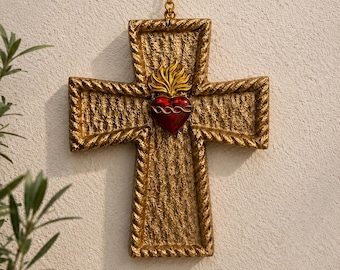 May include: A gold-colored cross-shaped ornament with a textured surface and rope-like edges. The center features a red heart with a gold flame design. A gold cord is attached for hanging. Religious decor.