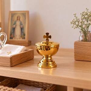 May include: A polished gold-tone metal ciborium featuring a rounded bowl, a pedestal base, and a lid topped with a small cross. The container has a reflective, smooth surface and is centered in the frame, showcasing its metallic finish and structure.