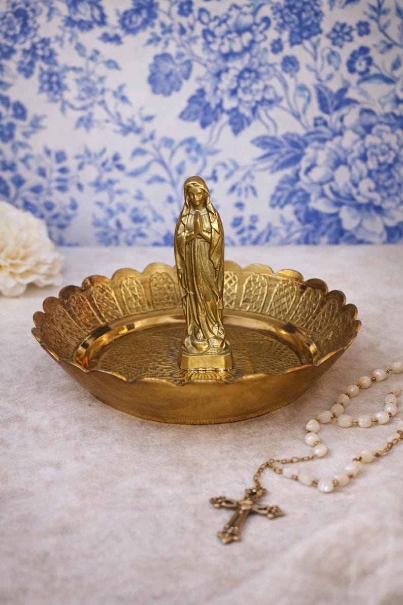 May include: A brass religious dish with a statue of the Virgin Mary. The dish has a scalloped edge and a detailed embossed design. A rosary with a cross is in the foreground. The background features a blue and white floral pattern.