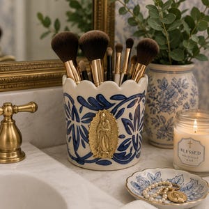 May include: A ceramic makeup brush holder with a blue and white floral design and a gold emblem. The holder is filled with various makeup brushes. A small dish with a pearl necklace and a candle are also visible.