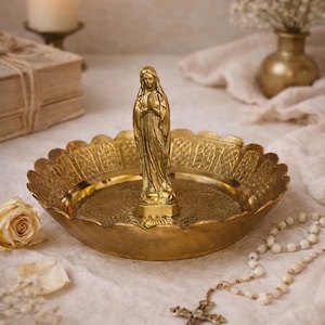 May include: A gold-colored brass religious display featuring a statue of a woman with clasped hands, set in a decorative dish. A rosary, a candle, and a vase of flowers are also present.