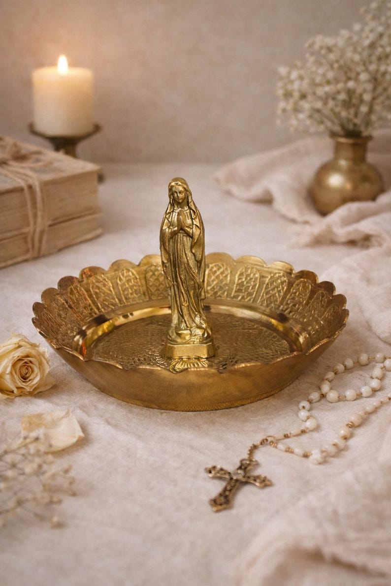 May include: A gold-colored brass religious display featuring a statue of a woman with clasped hands, set in a decorative dish. A rosary, a candle, and a vase of flowers are also present.