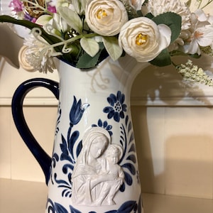 May include: A white ceramic jug with a dark blue handle and blue floral patterns. A raised white relief sculpture of a figure holding a child is centred on the front. The jug is filled with a bouquet of white artificial flowers.