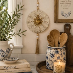 May include: A circular wall hanging featuring a textured cream-colored disc with a central gold-toned Sacred Heart emblem. The piece is suspended by a jute cord adorned with alternating natural wood and white beads, finished with a long jute tassel.