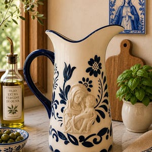 May include: A white ceramic pitcher with a blue handle and floral design. The pitcher features a bas-relief of Mary and baby Jesus. A small bowl of olives, a bottle of olive oil, and a religious icon are in the background.