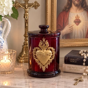 Puede incluir: Un frasco cilíndrico de vidrio rojo oscuro con tapa, que presenta un prominente emblema dorado y ornamentado de un corazón sagrado en el frente. El frasco tiene una superficie lisa y reflectante con un pequeño pomo en la tapa.