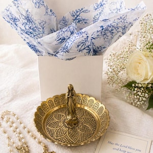 May include: A gold-colored decorative dish with a statue of a woman, a rosary, and a cross. The dish is round with a floral design. A white box with blue floral tissue paper is in the background. A card reads "May the Lord bless you and keep you."