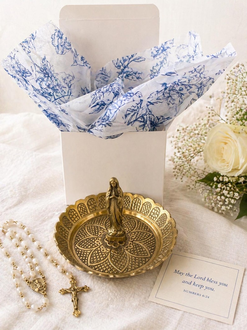 May include: A gold-colored decorative dish with a statue of a woman, a rosary, and a cross. The dish is round with a floral design. A white box with blue floral tissue paper is in the background. A card reads "May the Lord bless you and keep you."