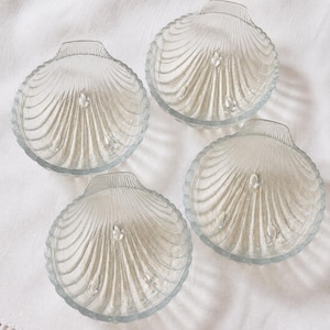 May include: A set of four clear glass dishes shaped like scallop shells. Each dish features a ribbed, fan-like pattern radiating from the hinge and is supported by three small, rounded glass feet on the underside for stability and elevation.