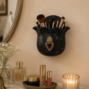 May include: A black wall-mounted holder with a scalloped rim and a gold-toned sacred heart emblem featuring a red center. The holder contains several makeup brushes with brown and black bristles, positioned above a vanity with various beauty products.