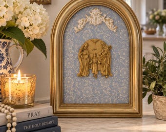 May include: An arched gold-toned frame containing a light blue floral patterned background. Centered is a gold-colored relief sculpture of the Holy Family, featuring Mary, Joseph, and the child Jesus, topped with a decorative white filigree ornament.
