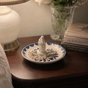 May include: A white ceramic dish with a blue floral design holds a white statue and a pearl necklace. The dish sits on a dark wooden nightstand next to a lamp and a vase of white roses.