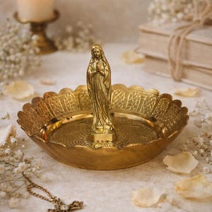 May include: A gold-colored religious trinket dish with a statue of the Virgin Mary. The dish has a scalloped edge and a detailed pattern. A gold cross necklace lies nearby. A lit candle and books are in the background.