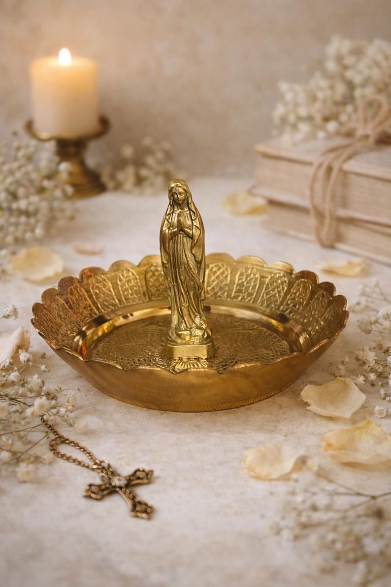 May include: A gold-colored religious trinket dish with a statue of the Virgin Mary. The dish has a scalloped edge and a detailed pattern. A gold cross necklace lies nearby. A lit candle and books are in the background.