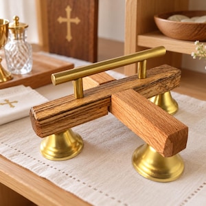 May include: A liturgical altar bell set featuring three brass bells mounted beneath a T-shaped wooden handle. The natural wood crossbar is topped with a horizontal brass handle, and the three bells are finished in a polished gold-tone metal.