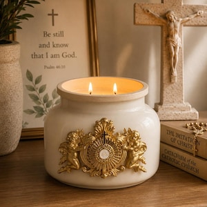 May include: A white ceramic candle jar with two lit wicks, featuring a gold-toned relief emblem on the front. The emblem depicts two cherubs flanking a central sunburst design with a small cross and the letters JHS inscribed in the center.
