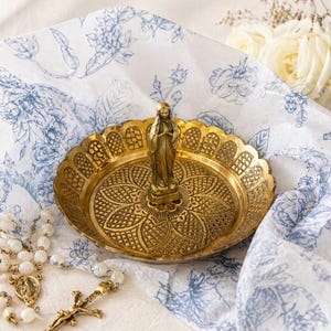 May include: A gold-colored decorative dish with a detailed floral pattern and a small statue of a woman. The dish is on a white and blue floral patterned cloth, with a rosary and a crucifix visible.