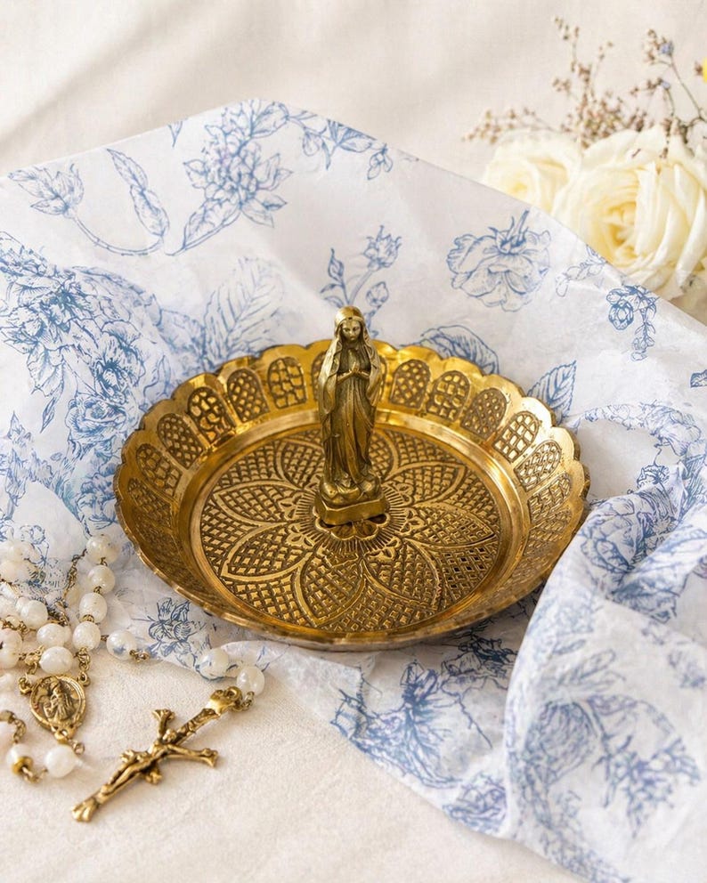 May include: A gold-colored decorative dish with a detailed floral pattern and a small statue of a woman. The dish is on a white and blue floral patterned cloth, with a rosary and a crucifix visible.
