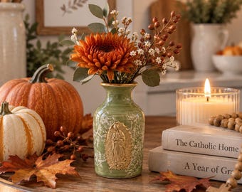 May include: A light green ceramic vase featuring a gold-toned embossed image of the Virgin of Guadalupe on the front, filled with a bouquet of orange chrysanthemums, small white flowers, and brown berries, set on a wooden surface.