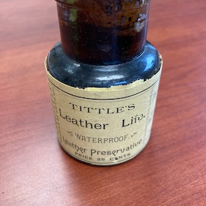以下が含まれることがあります： 赤い蓋と紙のラベルが付いた、小さな濃い茶色のガラス瓶。ラベルには「TITTLE'S Leather Life, WATERPROOF, Leather Preservative, PRICE 25 CENTS」と書いてあります。