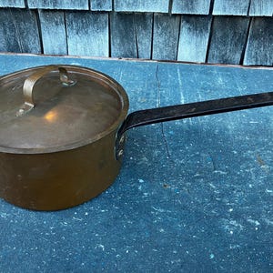 May include: A copper pot with a lid and a black handle. The pot is sitting on a blue surface.