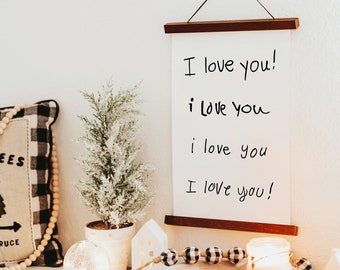 Puede incluir: Un lienzo blanco con el texto "I love you!" escrito cuatro veces en marcador negro, colgado de una varilla de madera. El lienzo está rodeado de elementos decorativos, incluyendo una almohada con un patrón de cuadros negros y blancos, un pequeño árbol en maceta y una casa de cerámica blanca.