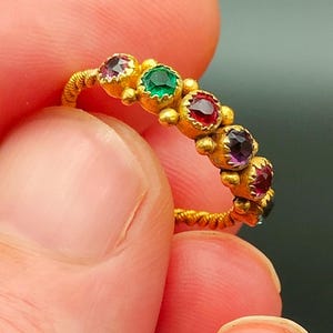 Puede incluir: Un anillo de oro con una banda retorcida y una fila de piedras preciosas de colores. Las piedras son rojas, verdes, moradas y rosas.