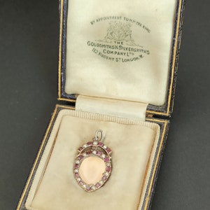 Puede incluir: Un medallón de oro con un borde de piedras rojas y blancas, en una caja de terciopelo negro y crema. La caja tiene la inscripción "BY APPOINTMENT TO HM THE KING THE GOLDSMITHS & SILVERSMITHS COMPANY LTD 112 REGENT ST LONDON.W."