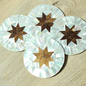 Può includere: Quattro sottobicchieri rotondi con un design a mosaico di madreperla. Ogni sottobicchiere presenta una stella centrale realizzata con guscio di cocco marrone. I sottobicchieri sono disposti su una superficie di legno chiaro.