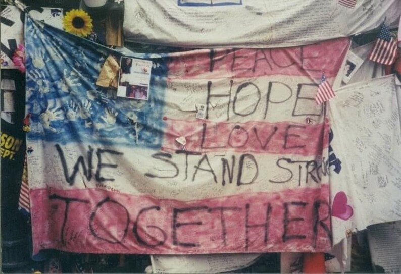 May include: A painted American flag with the words "PEACE HOPE LOVE WE STAND STRONG TOGETHER" and handprints. Additional flags and memorial items are visible in the background.