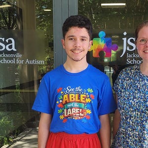 Może przedstawiać: Niebieski t-shirt z napisem See the ABLE not the LABEL kolorowymi literami w otoczeniu elementów układanki, noszony z czerwonymi szortami. W tle szklane drzwi z logo Jacksonville School for Autism i kolorowymi ikonami puzzli.