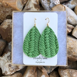 May include: A pair of green leather teardrop earrings with a woven pattern. The earrings are displayed in a white box with the text "DesignByTammi" and "designbytammi.store".