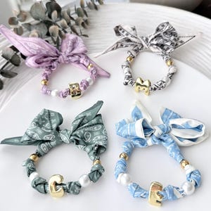 May include: Four hair scrunchies featuring fabric bows with paisley patterns in lavender, black and white, sage green, and light blue. Each scrunchie includes gold-toned metal beads, white faux pearls, and a single gold-toned letter charm.
