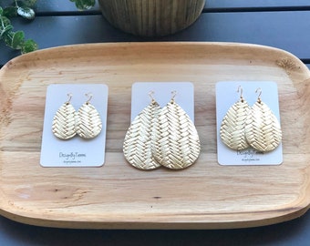 May include: Three pairs of gold leather earrings with a woven pattern. The earrings are displayed on a wooden tray. The earrings are all different sizes.