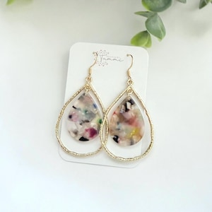 May include: A pair of gold-toned drop earrings. Each earring features a teardrop-shaped, multi-colored pendant within a textured gold hoop. The pendant has a mix of colors including pink, white, and black.
